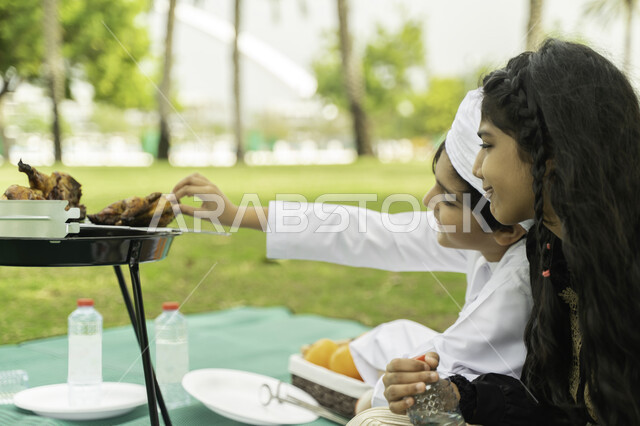 الحوارات والنقاش بين الاخوة، انشطة صيفيه ترفيهيه, صورة من الجانب لولد عربي اماراتي خليجي يرتدي الكندورة و العمامة البيضاء في نزهه في الهواء الطلق مع شقيقته في إحدى المنتزهات والحدائق الغناء، ولد وبنت سعوديان في الخارج يستمتعان بتحضير وجبة الغداء