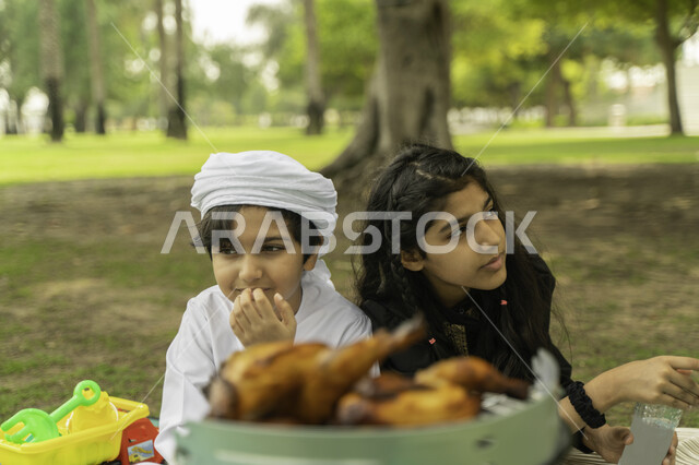 الاستمتاع بتحضير وجبة الغداء، انشطة صيفيه ترفيهيه, حفلة شواء في الخارج، صورة مقربة لولد عربي اماراتي خليجي يرتدي الكندورة و العمامة البيضاء  في نزهه في الهواء الطلق مع شقيقته في إحدى المنتزهات والحدائق الغناء، ولد وبنت سعوديان في الخارج ينتظران الدجاج الم