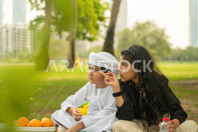 الاشارة الى شيء ما, نزهه في الهواء الطلق, صورة مقربة لولد عربي اماراتي خليجي يرتدي الكندورة و العمامة البيضاء يستمتع بقضاء وقت ممتع مع شقيقته في إحدى المنتزهات ، ولد وبنت سعوديان في الخارج, تبادل الاحاديث والحوارات بين الاشقاء