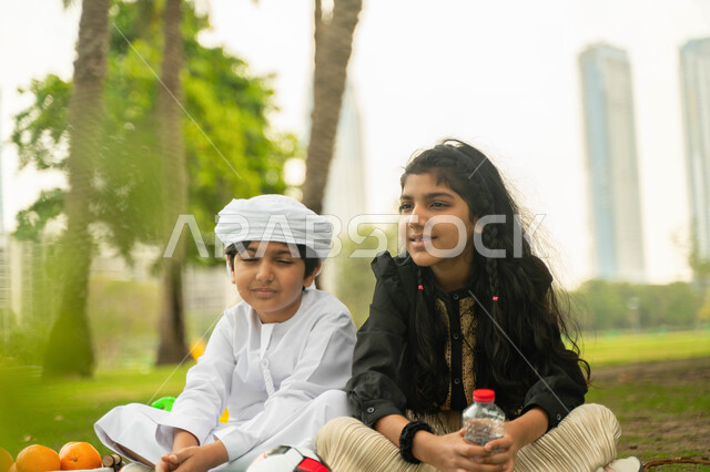 تبادل الاحاديث والحوارات بين الاشقاء، نزهه في الهواء الطلق, صورة مقربة لولد عربي اماراتي خليجي يرتدي الكندورة و العمامة البيضاء يستمتع بقضاء وقت ممتع مع شقيقته في إحدى المنتزهات والحدائق، ولد وبنت سعوديان في الخارج، مفهوم العلاقات الودية بين الاخوة