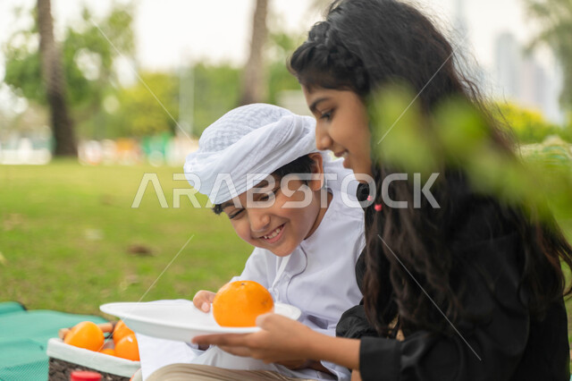 مفهوم العلاقات الودية بين الاخوة، صورة مقربة لولد عربي اماراتي خليجي يرتدي الكندورة و العمامة البيضاء يستمتع بقضاء وقت ممتع مع شقيقته في إحدى المنتزهات ، ولد وبنت سعوديان يقومان بمشاركة الفواكه الطازجة اللذيذة ، نزهه في الهواء الطلق