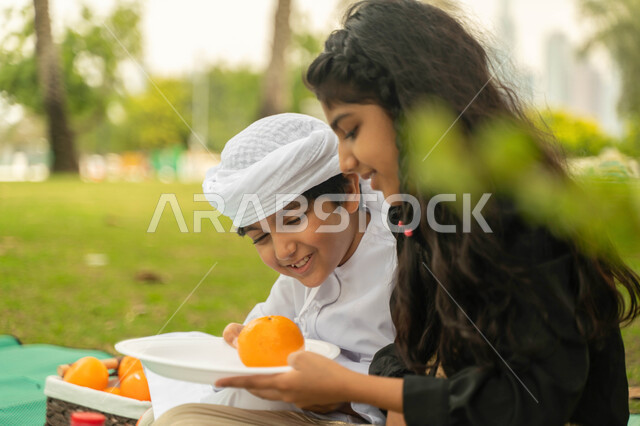 نزهه في الهواء الطلق, مفهوم العلاقات الودية بين الاخوة، صورة مقربة لولد عربي اماراتي خليجي يرتدي الكندورة و العمامة البيضاء يستمتع بقضاء وقت ممتع مع شقيقته في إحدى الحدائق، ولد وبنت سعوديان يقومان بمشاركة الفواكه الطازجة اللذيذة