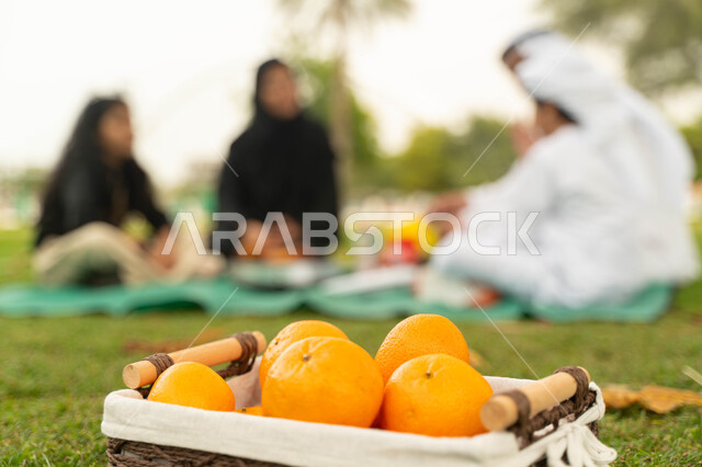 تناول الفواكه المنعشة واللذيذه, صورة مقربة لسلة خشبية مليئة بالبرتقال الطازج على العشب الاخضر, عائلة عربية اماراتية خليجية في إحدى المنتزهات في الخارج, عائلة سعودية تقضي أوقات ممتعه مع الابناء, منتجات وطنية سعودية محلية، سناك صحي ومفيد