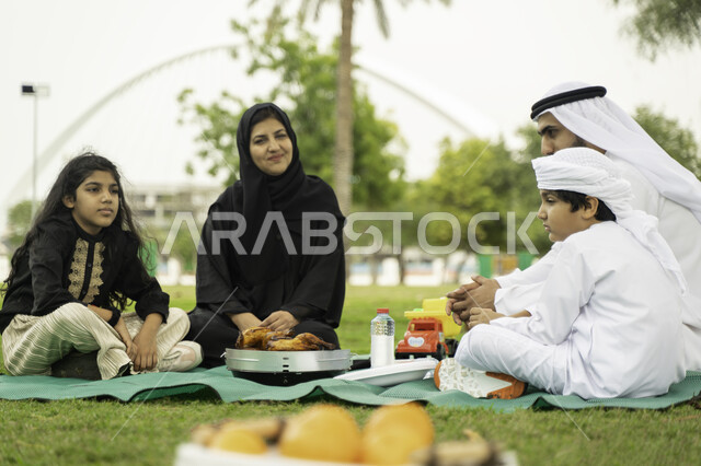 Distinctive family atmosphere, having lunch outside, a close-up of an Arab Emirati Gulf family sitting on a green rug, a Saudi family in one of the parks and gardens, spending quality time with children, looking at the camera, gestures indicating happines