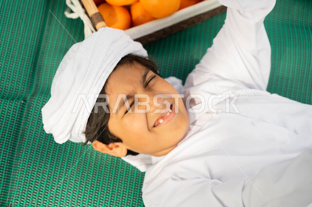 نزهة في الهواء الطلق, اللعب تحت اشعه الشمس في أجواء صيفيه، صورة مقربة من الاعلى لولد عربي إماراتي خليجي يرتدي الكندوره والعمامه البيضاء بجانبه سلة من البرتقال, طفل سعودي يستلقي على بساط اخضر في الخارج، الاهتمام بالاطفال والاعتناء بهم