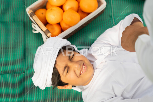 اللعب تحت اشعه الشمس في أجواء صيفيه، قضاء أوقات ممتعة مع الابناء, صورة مقربة من الاعلى لولد عربي إماراتي خليجي يرتدي الكندوره والعمامه البيضاء بجانبه سلة من البرتقال, طفل سعودي يستلقي على بساط اخضر في الخارج، الاهتمام بالاطفال والاعتناء بهم