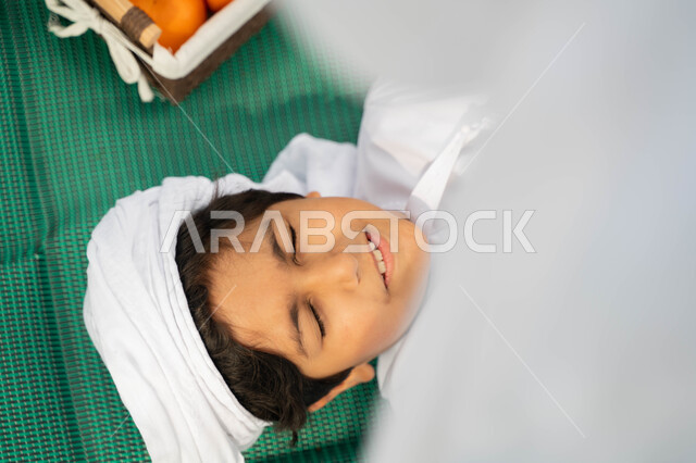 الاعتناء بالاطفال والاهتمام بهم، الاسترخاء تحت اشعه الشمس في  أجواء صيفيه، قضاء أوقات ممتعة باللعب مع الابناء, صورة مقربة من الاعلى لولد عربي إماراتي خليجي يرتدي الكندوره والعمامه البيضاء بجانبه سلة من البرتقال, طفل سعودي يستلقي على بساط اخضر في الخارج