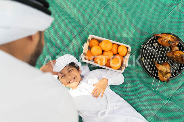 الاهتمام بالاطفال والاعتناء بهم، ايماءات تدل على السعادة، قضاء أوقات ممتعة باللعب والتنزه مع الابناء, صورة من الاعلى لولد عربي إماراتي خليجي يرتدي الكندوره والعمامه الييضاء بجانبه سلة من البرتقال والدجاج المشوي, طفل سعودي يستلقي على بساط اخضر في الخارج