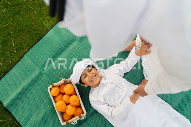 ايماءات تدل على السعادة، اللعب مع الاطفال والاعتناء بهم، قضاء أوقات ممتعة باللعب والتنزه مع الابناء, صورة من الاعلى لولد عربي إماراتي خليجي يرتدي الكندوره والعمامه الييضاء بجانبه سلة من البرتقال الطازج, طفل سعودي يستلقي على بساط اخضر في احدى المنتزهات