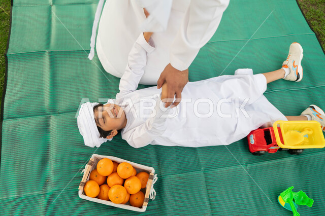 اللعب مع الاطفال والاعتناء بهم، قضاء أوقات ممتعة باللعب والتنزه مع الابناء, صورة من الاعلى لولد عربي إماراتي خليجي يرتدي الكندوره والعمامه الييضاء بجانبه سلة من البرتقال الطازج, طفل سعودي يستلقي على بساط اخضر في احدى المنتزهات ، انشطة ترفيهيه صيفيه