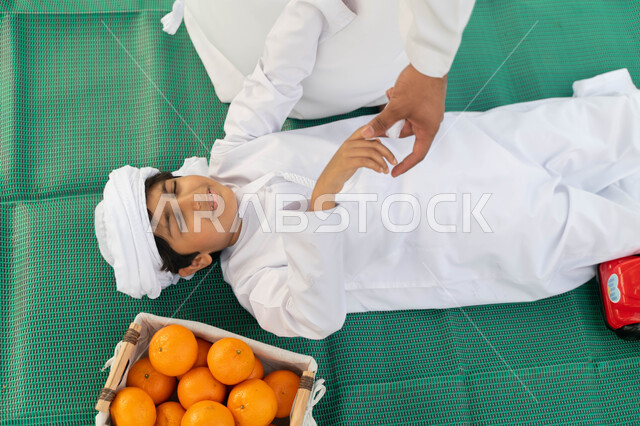 قضاء أوقات ممتعة باللعب والتنزه مع الابناء, صورة من الاعلى لولد عربي إماراتي خليجي يرتدي الكندوره والعمامه الييضاء بجانبه سلة من البرتقال الطازج, طفل سعودي يستلقي على بساط اخضر في احدى المنتزهات ، اللعب مع الاطفال والاعتناء بهم، انشطة ترفيهيه صيفيه
