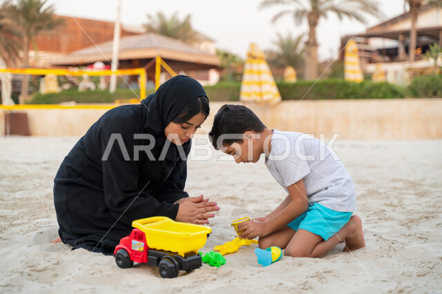 إيماءات التركيز في اللعب برمال البحر الناعمة,  تغيير الاجواء المنزلية وزيادة الترابط الاسري بين الابناء والاهل، السياحة في دول الخليج العربي والاستمتاع بجمال الرمال وبناء المجسمات الرملية, امرأة عربية خليجية سعودية تقضي وقت ممتع مع إبنها على ساحل البحر