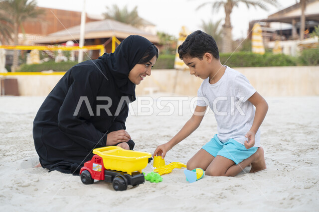 إيماءات وجه تدل على السعادة, تغيير الاجواء المنزلية وزيادة الترابط الاسري بين الابناء والاهل، السياحة في دول الخليج العربي والاستمتاع بجمال الرمال, امرأة عربية خليجية سعودية تقضي وقت ممتع مع إبنها على ساحل البحر, اللعب بادوات الرمل وبناء المجسمات الرملية