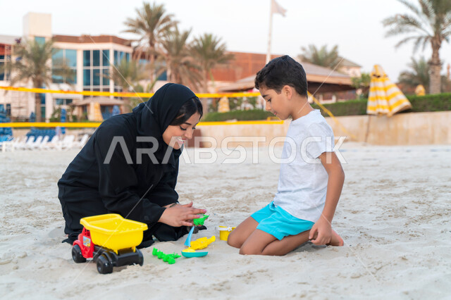 Tourism in the Arab Gulf countries and enjoying the summer vacation, a smiling Saudi Gulf Arab woman spending quality time with her son on the seashore, the concept of a happy, interconnected Saudi family, family entertainment activities, developing the c