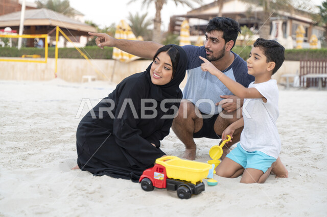 قضاء وقت ممتع مع الابناء, مفهوم الاسرة السعودية السعيدة المترابطة، السياحة في دول الخليج العربي والاستمتاع بالاجازة الصيفية، الاشارة والنظر الى شيء ما، عائلة عربية خليجية اماراتية في عطلة صيفية تجلس وتلعب على رمال شاطئ البحر، نشاطات ترفيهية عائلية