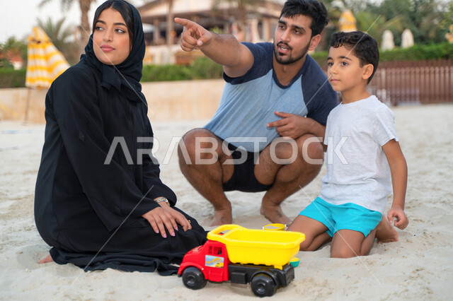 الاشارة والنظر الى شيء ما، قضاء وقت ممتع مع الابناء, عائلة عربية خليجية اماراتية في عطلة صيفية تجلس وتلعب على رمال شاطئ البحر، السياحة في دول الخليج العربي والاستمتاع بالاجازة، مفهوم الاسرة السعودية السعيدة المترابطة، نشاطات ترفيهية عائلية