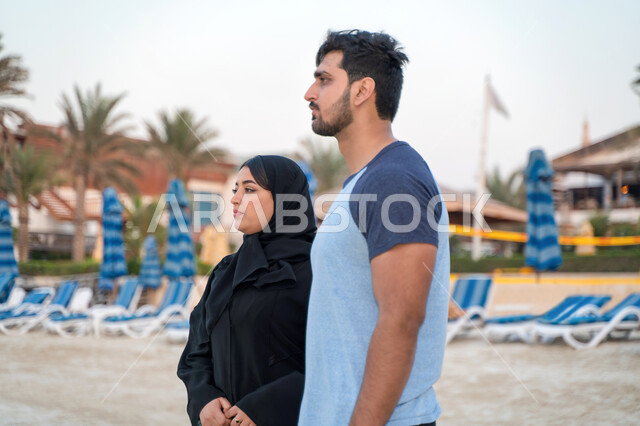 مفهوم الاسرة السعودية السعيدة، ايماءات النظر الى شيء ما، صورة من الجانب لزوجان عربيان خليجيان اماراتيان يقفان على شاطئ البحر، السياحة في دول الخليج العربي والاستمتاع بالاجازة الصيفية، الإستجمام والتنزه في الهواء الطلق, رجل سعودي وزوجته يقضيان عطلة صيفية