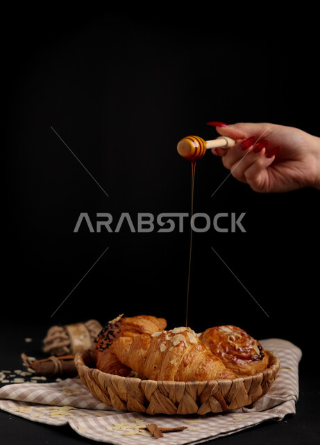 يد مرأة تضع العسل الطبيعي على الكرواسون ، وجبة فطور عربية مصورة على خلفية سوداء ، معجنات و مخبوزات عربية ، مأكولات و وجبات صحية ، فطور صحي ولذيذ ، كرواسون ، ملعقة العسل ، غذاء صحي
