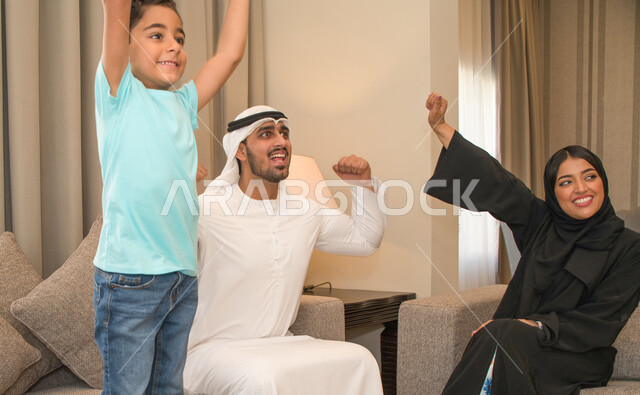 قضاء أوقات عائليه ممتعة، ايماءات وجه ويدين تدل على الحماس والفرحة بالإنتصار، عائله عربيه خليجيه اماراتيه تقوم بمشاهدة المباراة عبر التلفاز، أسرة سعوديه تجلس في غرفه المعيشه، شغف تشجيع فريق كره القدم المفضل، متابعه المنتخب الوطني الاماراتي.