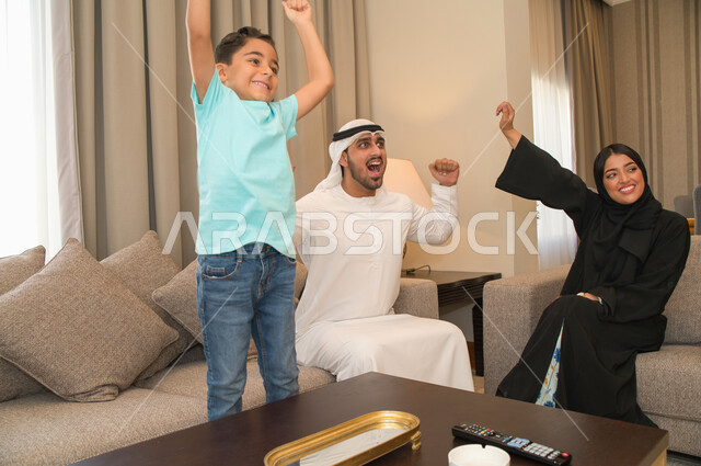 ايماءات وجه ويدين تدل على الحماس والفرحة بالإنتصار، عائله عربيه خليجيه اماراتيه تقوم بمشاهدة المباراة عبر التلفاز، أسرة سعوديه تجلس في غرفه المعيشه، قضاء أوقات عائليه ممتعة، شغف تشجيع فريق كره القدم المفضل، متابعه المنتخب الوطني الاماراتي.