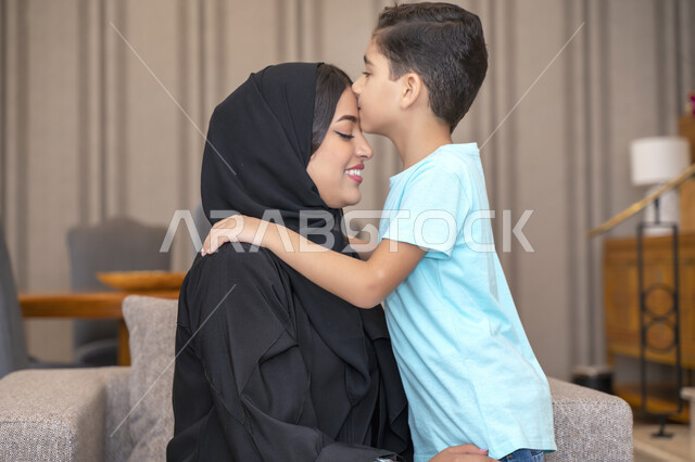 Gestures indicating love and tenderness, the role of the mother in the good upbringing of her children, an Arab Emirati boy kissing his mother’s head, a smiling Saudi Arab woman wearing a black Gulf abaya, the concept of intimacy and family bonding, the rights of mothers over children.