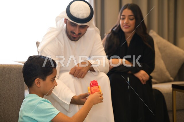 تخصيص وقت من الأهل لممارسه النشاطات الترفيهية مع الأولاد، أسرة عربيه خليجيه اماراتيه تستمتع بقضاء أوقات الفراغ معا، ولد سعودي يجلس في غرفه المعيشه يقوم بتركيب المكعبات بمشاركه عائلته، المحافظه على الترابط الأسري والاستقرار العائلي.