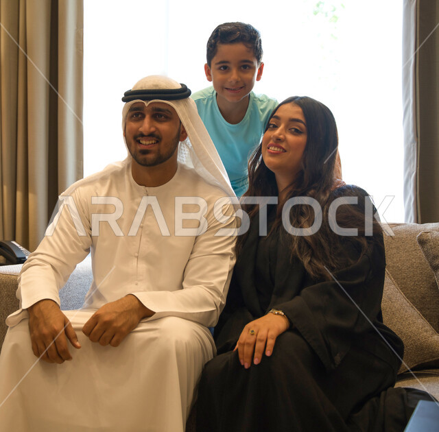 Love and intimacy between fathers and children, the concept of family security and stability, a happy family atmosphere full of love and tenderness, an Emirati Arab family sitting in the living room, a Saudi family looking at the camera, gestures of joy and happiness to spend quality time with the family.