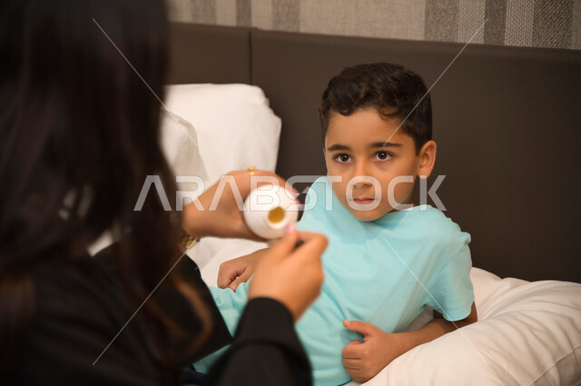 معاناة الأطفال أثناء مرضهم واهتمام الأم بأولادها وتأثيره في حياة الأبناء, الالتزام بالأوقات الصحيحة لتناول الدواء وأخذه بصورة آمنة, صورة مقربة لولد عربي خليجي إماراتي يمر بحالة من المرض وينظر لوالدته, أم سعودية تعطي ابنها المريض الشراب المناسب
