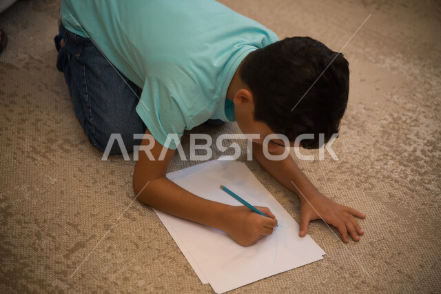 ممارسه الهوايات والأنشطه الترفيهية المفضله، قضاء أوقات ممتعه في المنزل، تنمية المهارات الفكريه والذهنيه لدى الأطفال، صبي عربي خليجي اماراتي يقوم بالرسم والتلوين، طفل سعودي يجلس على السجاده، استغلال وقت الفراغ في التعليم والتدريب.