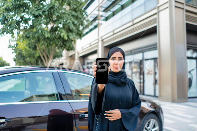خدمات توصيل الركاب الآمنة، مفهوم شراء وأجار السيارات الحديثة عن طريق الاونلاين، طلب سيارة تاكسي بواسطة الجوال، عرض شاشة سواداء على الهاتف المحمول، إمرأة عربية اماراتية خليجية ترتدي الحجاب والعباءة السوداء، شابة سعودية تقف بجانب السيارة