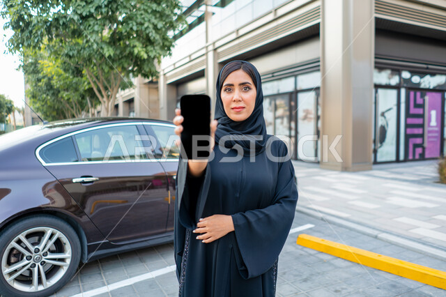 أجار و شراء السيارات الحديثة عن طريق الاونلاين، طلب سيارة تاكسي بواسطة الجوال، عرض شاشة سواداء على الهاتف المحمول، إمرأة عربية اماراتية خليجية ترتدي الحجاب والعباءة السوداء، شابة سعودية تقف بجانب السيارة، خدمات توصيل الركاب الآمنة