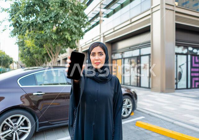 Buying and renting modern cars via the Internet, ordering a taxi via mobile phone, displaying a black screen on the mobile phone, an Arab Emirati Gulf woman wearing the hijab and black abaya, a young Saudi woman standing next to the car, safe passenger de