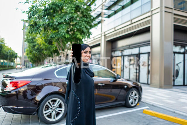 ايماءات وجه ويدين تدل عللى السعادة، مفهوم شراء وأجار السيارات الحديثة عن طريق الاونلاين، إمرأة عربية اماراتية خليجية ترتدي الحجاب والعباءة السوداء، طلب سيارة تاكسي بواسطة الجوال، شابة سعودية تقوم بعرض شاشة سواداء على الهاتف المحمول