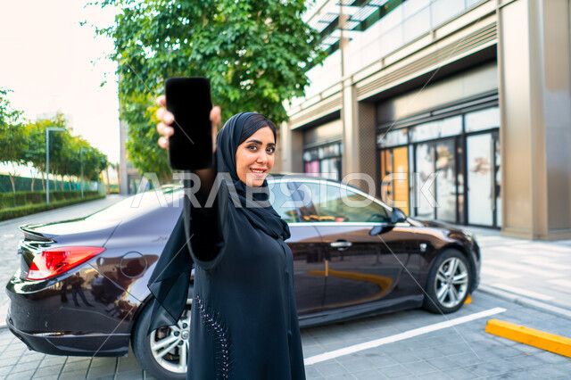 Face and hand gestures indicating happiness, the concept of buying and renting modern cars via the Internet, an Arab Emirati Gulf woman wearing the hijab and black abaya, ordering a taxi via mobile phone, a young Saudi woman displaying a black screen on h