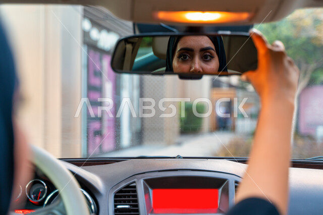 Adjusting the rear-view mirror of the car, the reflection of the image of a veiled Arab-Emirati-Gulf driver in the mirror of the car, the concept of independence, freedom and self-reliance, freedom of driving for Arab women, a Saudi woman wearing the blac
