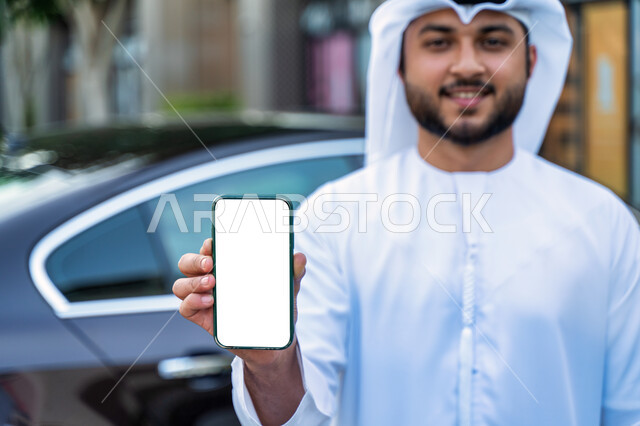 عرض شاشة بيضاء على الهاتف المحمول، صورة مقربة لرجل عربي اماراتي خليجي يرتدي الكندورة والغترة الإماراتية، شاب سعودي مبتسم يقف بجانب السيارة، مفهوم شراء وأجار السيارات الحديثة عن طريق الاونلاين، طلب سيارة تاكسي بواسطة الجوال