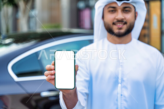 Requesting a taxi by mobile phone, displaying a white screen on the mobile phone, a close-up of an Arab Emirati Gulf man wearing an Emirati kandora and ghutra, a smiling Saudi young man standing next to the car, the concept of buying and renting modern ca