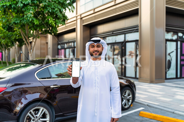 مفهوم شراء وأجار السيارات الحديثة عن طريق الاونلاين، عرض شاشة بيضاء على الهاتف المحمول، رجل عربي اماراتي خليجي يرتدي الكندورة والغترة الإماراتية, شاب سعودي مبتسم يقف بجانب السيارة، طلب سيارة تاكسي بواسطة الجوال