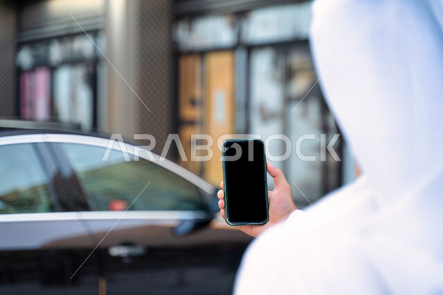 شراء وأجار السيارات عن طريق الاونلاين، طلب سيارة تاكسي بواسطة الجوال، عرض شاشة سواداء على الهاتف المحمول، صورة من الخلف لرجل عربي اماراتي خليجي يرتدي الكندورة والغترة الإماراتية، شاب سعودي يقف بجانب السيارة