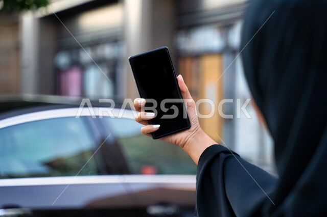 The concept of buying and renting modern cars via the Internet, ordering a taxi via mobile phone, displaying a black screen on the mobile phone, a picture from the back of an Arab Emirati Gulf woman wearing the hijab and black abaya, a young Saudi woman s