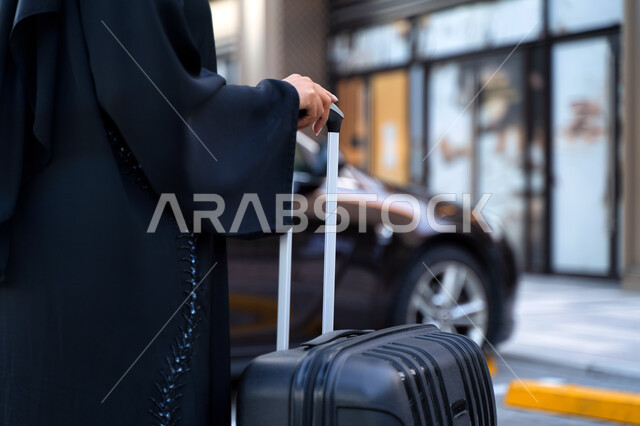 السياحة والسفر عن طريق البر، امرأة عربية خليجية إماراتية محجبة ترتدي العباءة السوداء تمسك بيدها حقيبة سفر، خدمة توصيل الركاب من وإلى المطار، شابة سعودية تتجه إلى السيارة، خدمات النقل المريحة و الآمنة، رحلات سياحية ممتعة