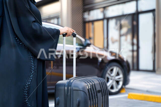  رحلات سياحية ممتعة، إمرأة عربية خليجية إماراتية محجبة ترتدي العباءة السوداء تمسك بيدها حقيبة سفر، خدمة توصيل الركاب من وإلى المطار، شابة سعودية تتجه إلى السيارة، خدمات النقل المريحة و الآمنة، مفهوم السياحة والسفر عن طريق البر