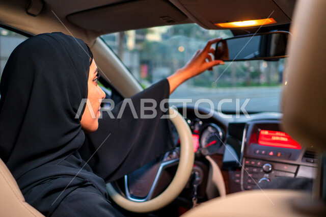 Driving with caution and following safety rules, the reflection of a veiled Arab Emirati driver in the car mirror, the concept of independence, freedom and self-reliance, freedom of driving for Arab women, a Saudi woman wearing a black abaya adjusting the