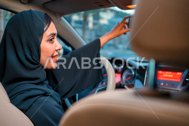 قيادة السيارة في شوارع مدينة دبي، صورة من الجانب لشابة عربية إماراتية خليجية محجبة مبتمسة، مفهوم الاستقلال و الحرية و الاعتماد على النفس، سيده سعودية ترتدي العباءة السوداء تقوم بتعديل مرآة الرؤية الخلفية في سيارتها