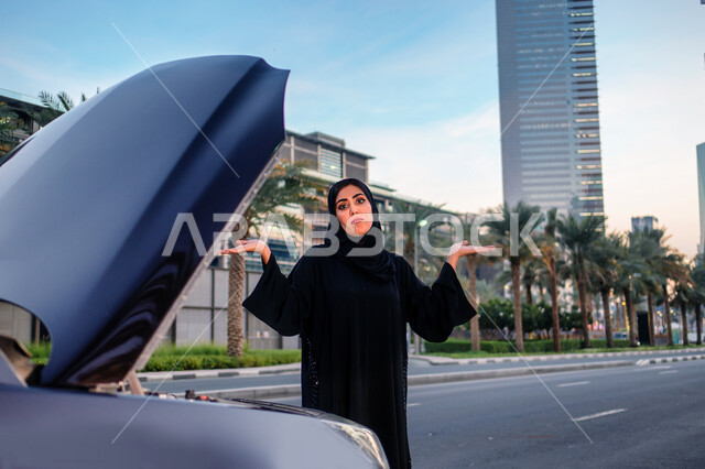 ايماءات يدين ووجه تدل على عدم معرفة العطل، مفهوم الاستقلال و الحرية و الاعتماد على النفس، إمرأه عربية خليجية إماراتيه محجبة تقف بالقرب من سيارتها المعطلة وغطاء المحرك مفتوح، سيده سعودية ترتدي العباءة السوداء، خلفية مجمع أبراج الإمارات