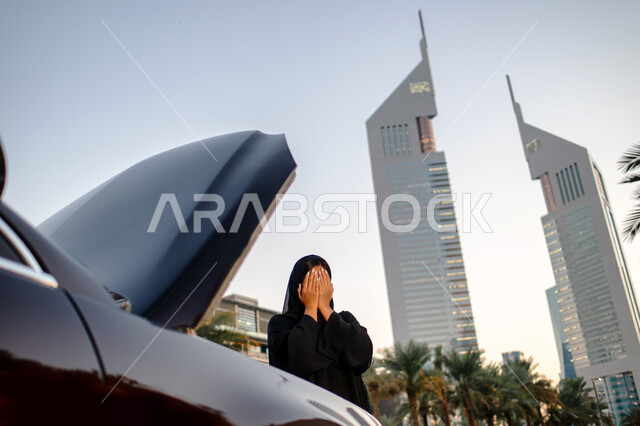 عدم معرفة العطل في السيارة، امرأة عربية خليجية إماراتية تقف أمام سيارتها المعطلة في الشارع وغطاء المحرك مفتوح، ايماءات البكاء والحزن لوقوع حادث سياره، شابة سعودية محجبة ترتدي العباءة السوداء، خلفية مجمع أبراج الإمارات
