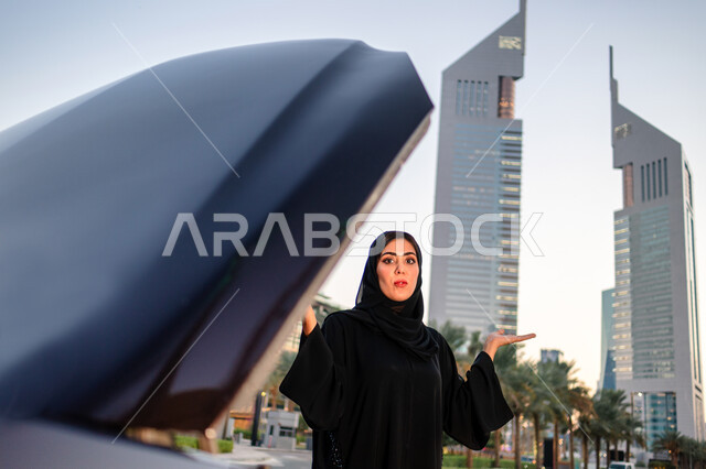 اعتماد المرأة على نفسها في التنقل والمواصلات، إمرأه عربية خليجية إماراتيه محجبة تقف بالقرب من سيارتها المعطلة وغطاء المحرك مفتوح، سيده سعودية ترتدي العباءة السوداء، ايماءات وجه ويدين تدل على عدم معرفة العطل، خلفية مجمع أبراج الإمارات