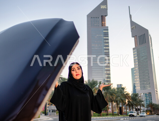Face and hand gestures indicating lack of knowledge of holidays, the concept of independence, freedom and self-reliance, a veiled Emirati Arab Gulf woman standing near her broken-down car with the hood open, a Saudi woman wearing a black abaya, the backgr
