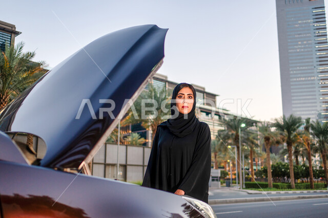 تحضير السيارة قبل القيادة، مفهوم الاستقلال و الحرية و الاعتماد على النفس، إمرأه عربية خليجية إماراتيه محجبة تقف بالقرب من سيارتها المعطلة وغطاء المحرك مفتوح، سيده سعودية ترتدي العباءة تنظر إلى الكاميرا، اعتماد المرأه على نفسها في التنقل و المواصلات