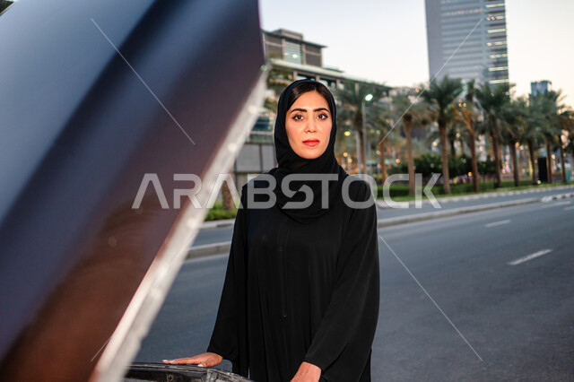 Independence, freedom and self-reliance, a veiled Emirati Arab Gulf woman standing near her broken car with the hood open, freedom of driving for Arab women, a Saudi woman wearing the abaya looking at the camera, women’s self-reliance in movement and tran